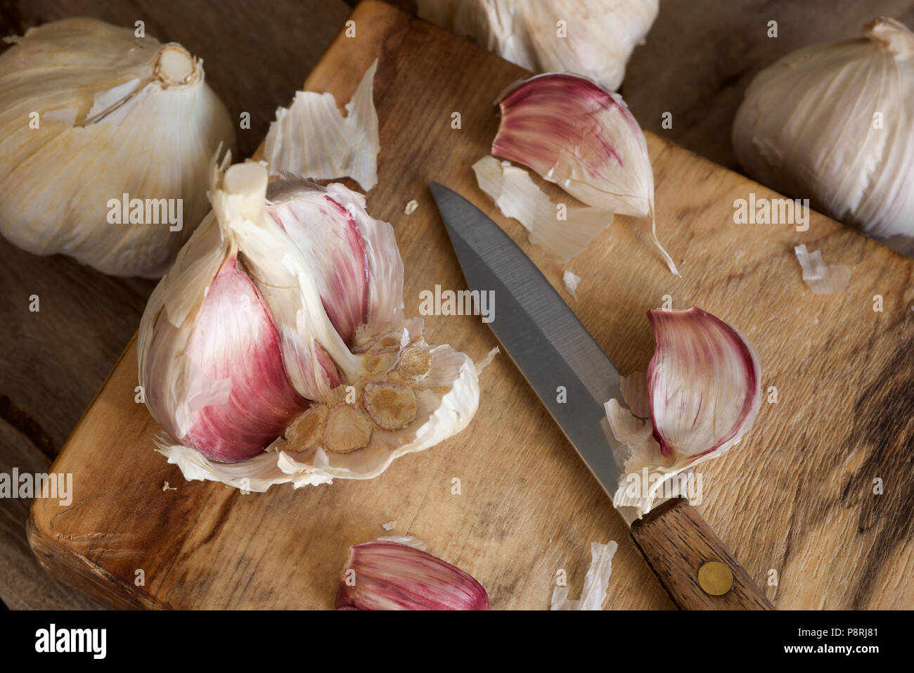 Viola organico bulbi di aglio e chiodi di garofano con una impugnatura in legno coltello seghettato essendo preparato su un tagliere di legno Foto Stock