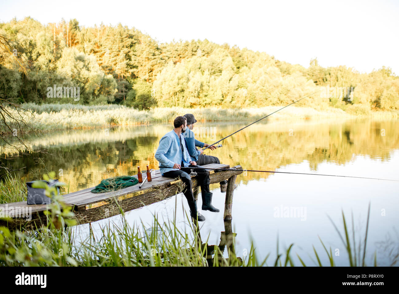 Due amici maschi vestiti di magliette blu insieme di pesca con rete e asta di seduta sul molo in legno durante la luce del mattino sul lago Foto Stock