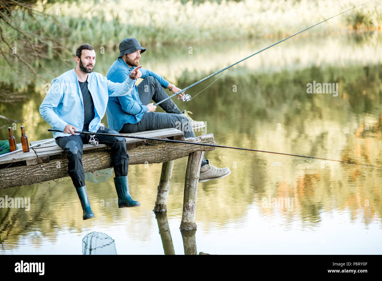 Due amici maschi vestiti di magliette blu insieme di pesca con rete e asta di seduta sul molo in legno durante la luce del mattino sul lago Foto Stock