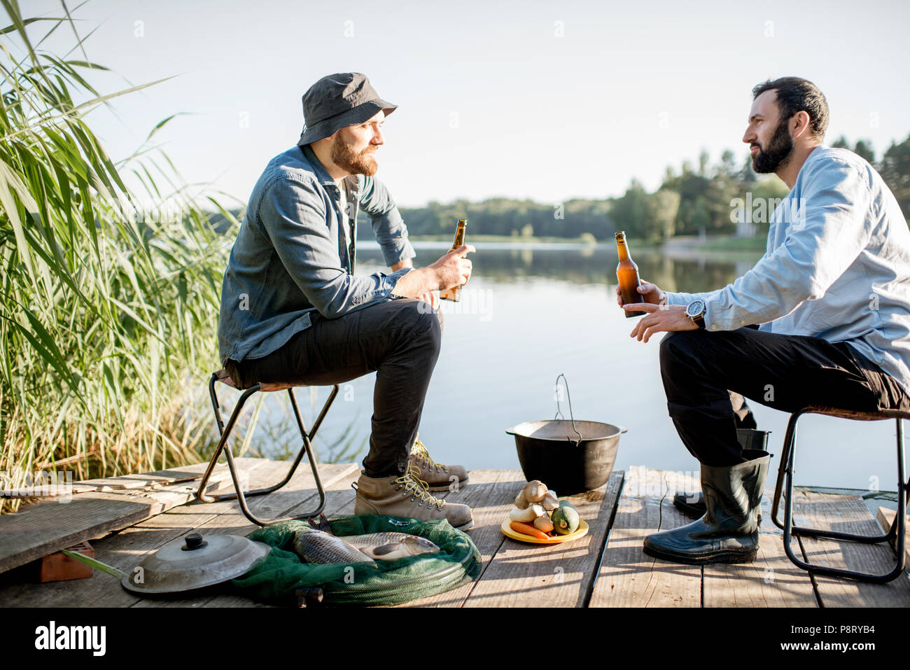 Due amici maschi rilassante con birra seduti insieme durante il processo di pesca sul molo vicino al lago Foto Stock