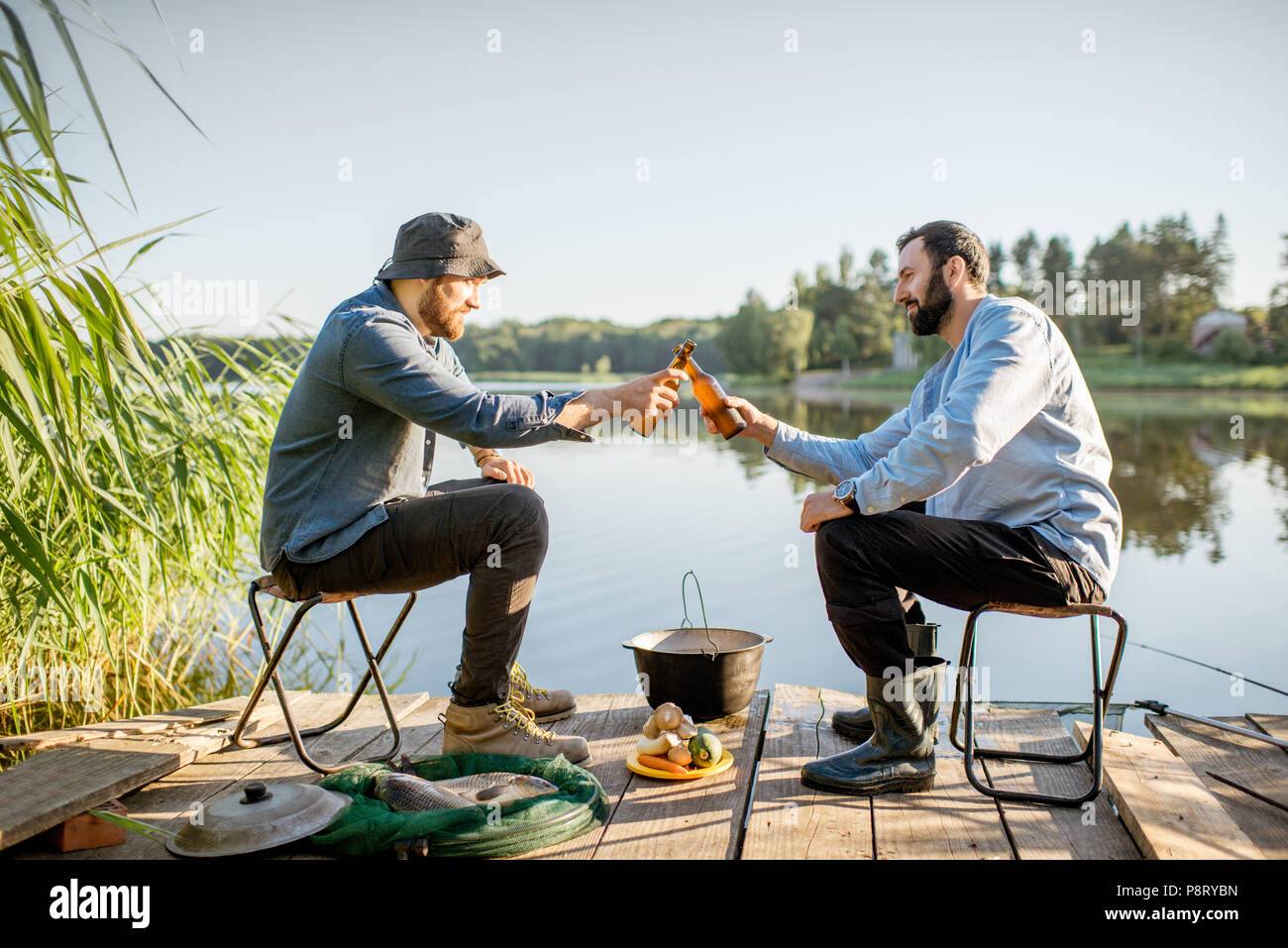 Due amici maschi tintinnio di bottiglie di birra seduti insieme durante il processo di pesca sul molo vicino al lago Foto Stock