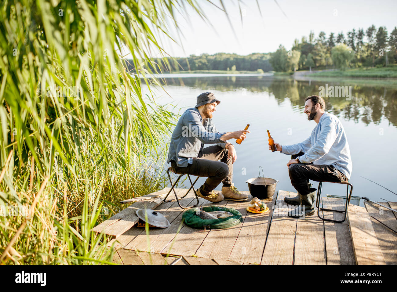 Due amici maschi rilassante con birra seduti insieme durante il processo di pesca sul molo vicino al lago Foto Stock