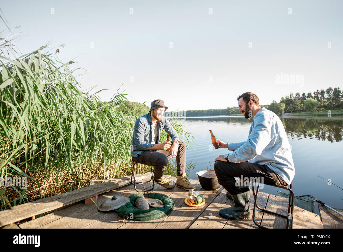 Panorama sul lago con due amici maschi seduti insieme con la birra durante il processo di pesca Foto Stock