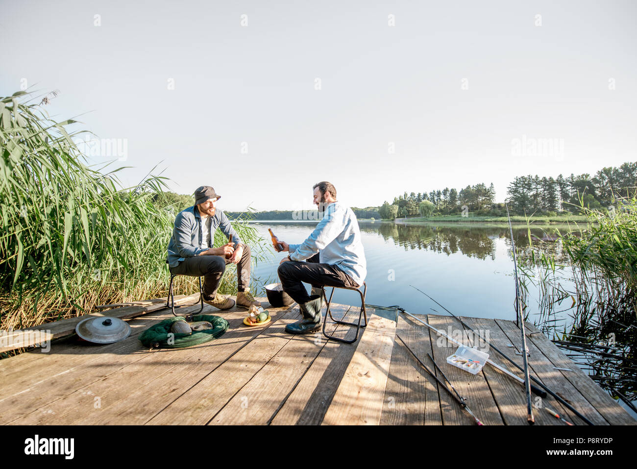 Panorama sul lago con due amici maschi seduti insieme con la birra durante il processo di pesca Foto Stock