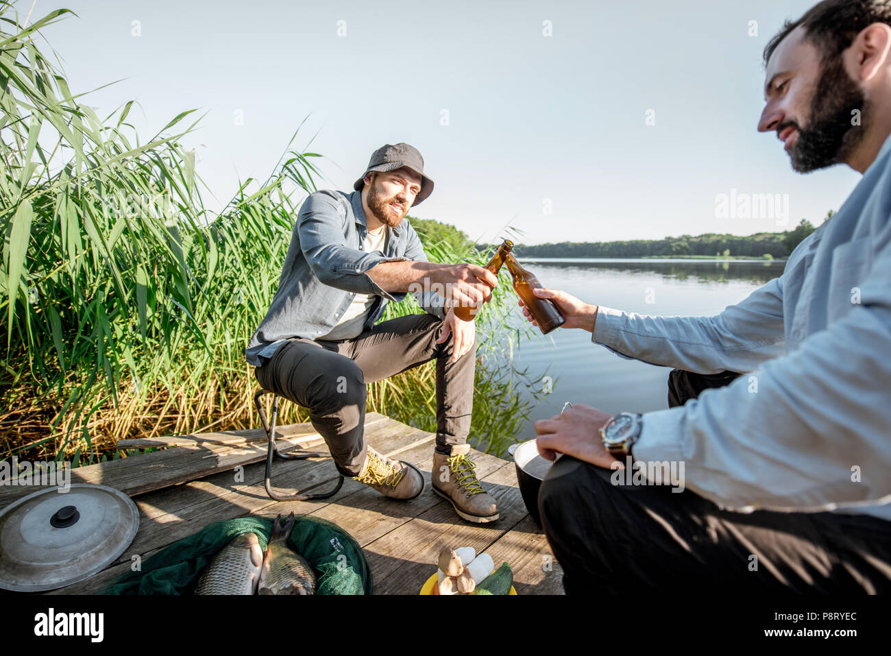 Due amici maschi tintinnio di bottiglie di birra seduti insieme durante il processo di pesca sul molo vicino al lago Foto Stock