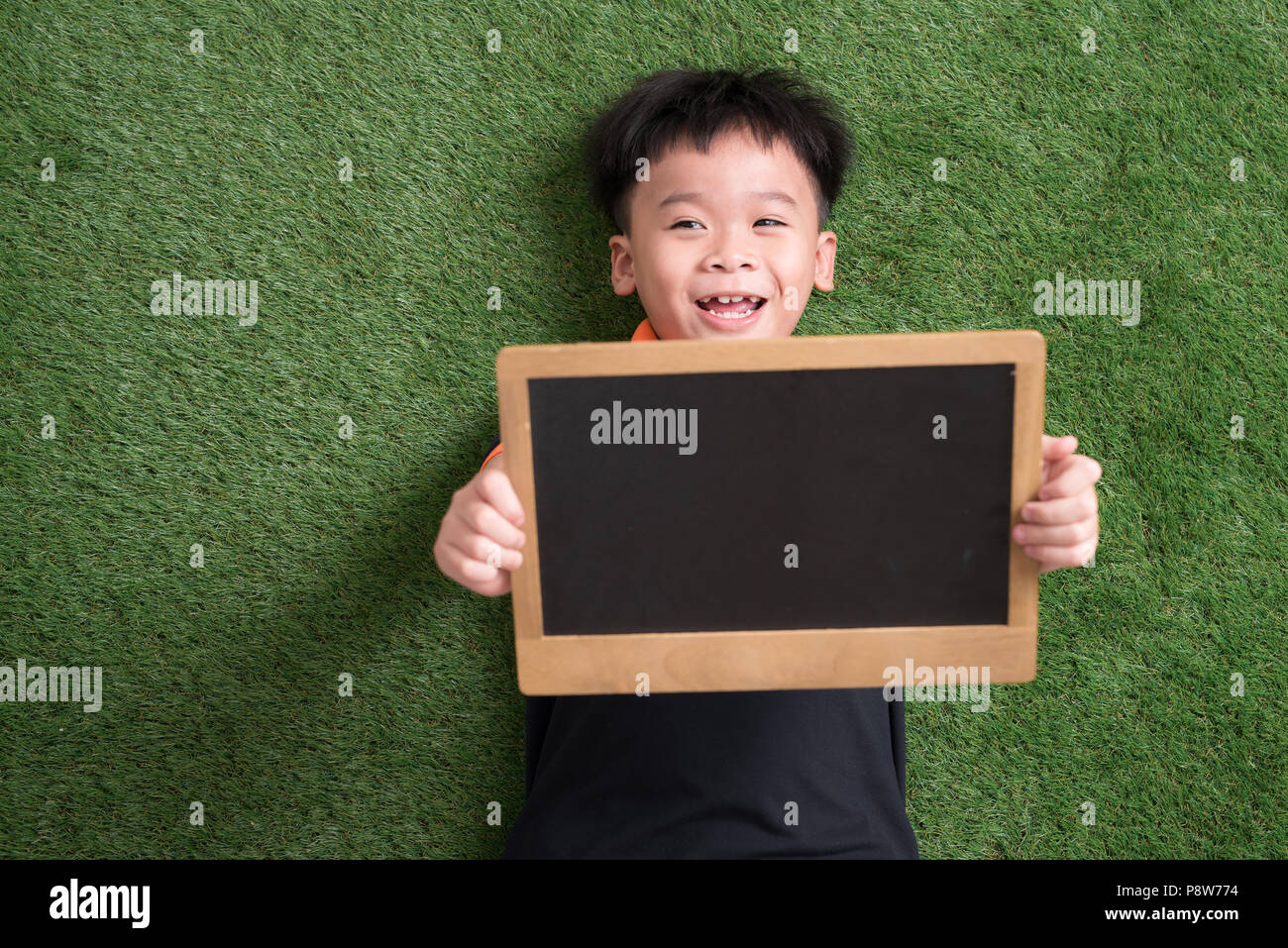 Carino asian bambino giacente e tenendo la lavagna su erba verde Foto Stock