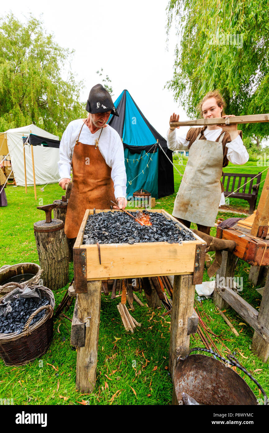 Storia viva, outdoor rievocazione evento. L uomo medievale, fabbro, utilizzando forge per rendere le armi di ferro, mentre il giovane apprendista aziona il mantice Foto Stock