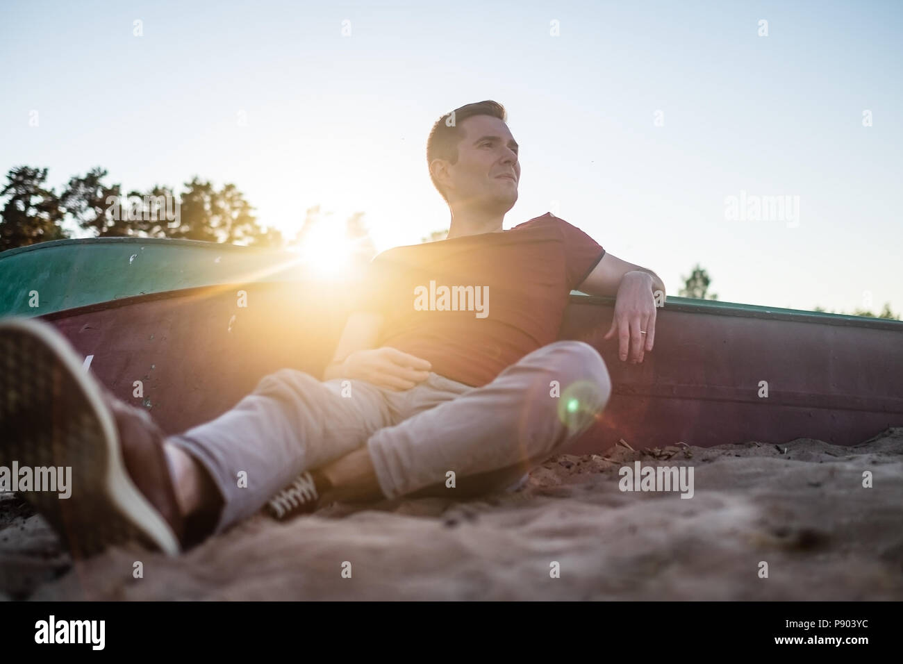 Uomo seduto sulla spiaggia di sabbia vicino a barca e guardando a parte su acqua di mare. Foto Stock
