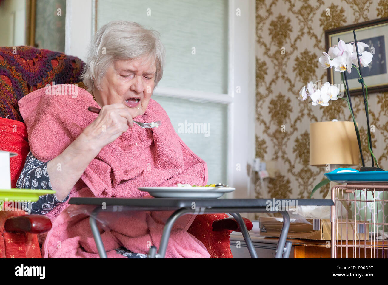 Old Lady con demenza seduto nella sua sedia reclinabile a mangiare il suo pranzo da una tabella con un asciugamano come un bib Foto Stock