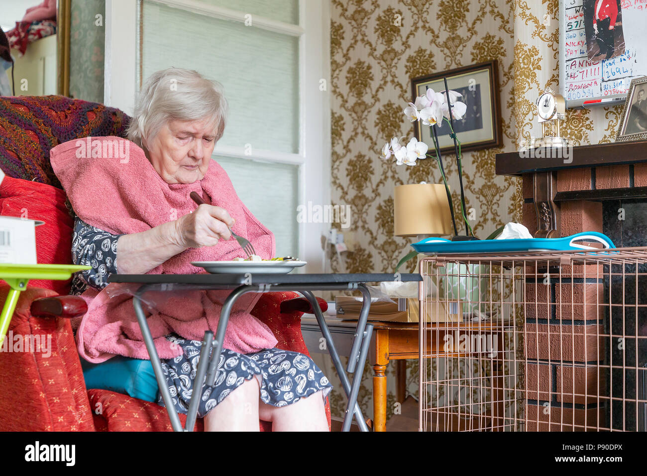 Old Lady con demenza seduto nella sua sedia reclinabile a mangiare il suo pranzo da una tabella con un asciugamano come un bib Foto Stock