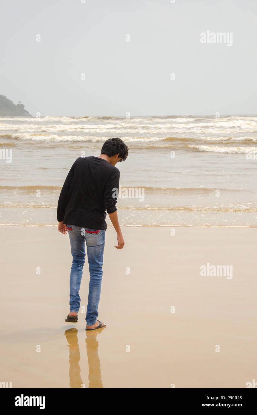 Vista dal retro di un ragazzo in abiti casual che guarda la sabbia bagnata in spiaggia Betul, Cavelossim, Goa, India Foto Stock