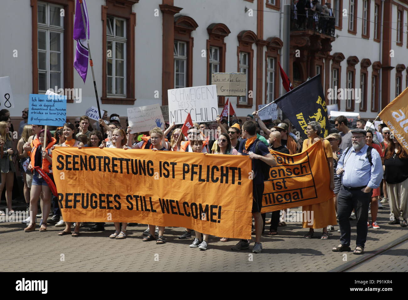 Mainz, Germania. Il 14 luglio 2018. Manifestanti marzo trogolo Mainz con striscioni e bandiere. Il banner principale legge 'Sea di salvataggio è obbligatorio - rifugiati ancora benvenuto". A poche centinaia di manifestanti hanno marciato attraverso Mainz, per protestare contro la politica del governo tedesco e della UE di chiusura delle frontiere e la prevenzione di mare le operazioni di soccorso delle ONG, mettendo a terra le loro navi a Malta, che fa sì che i rifugiati di affogare nel Mediterraneo. Hanno chiamato per il sindaco del comune di Magonza a prendere in rifugiati provenienti dal Mediterraneo. La protesta è stata parte della tedesca ampia Seebrucke (mare) ponte di protesta che ha visto thousan Foto Stock