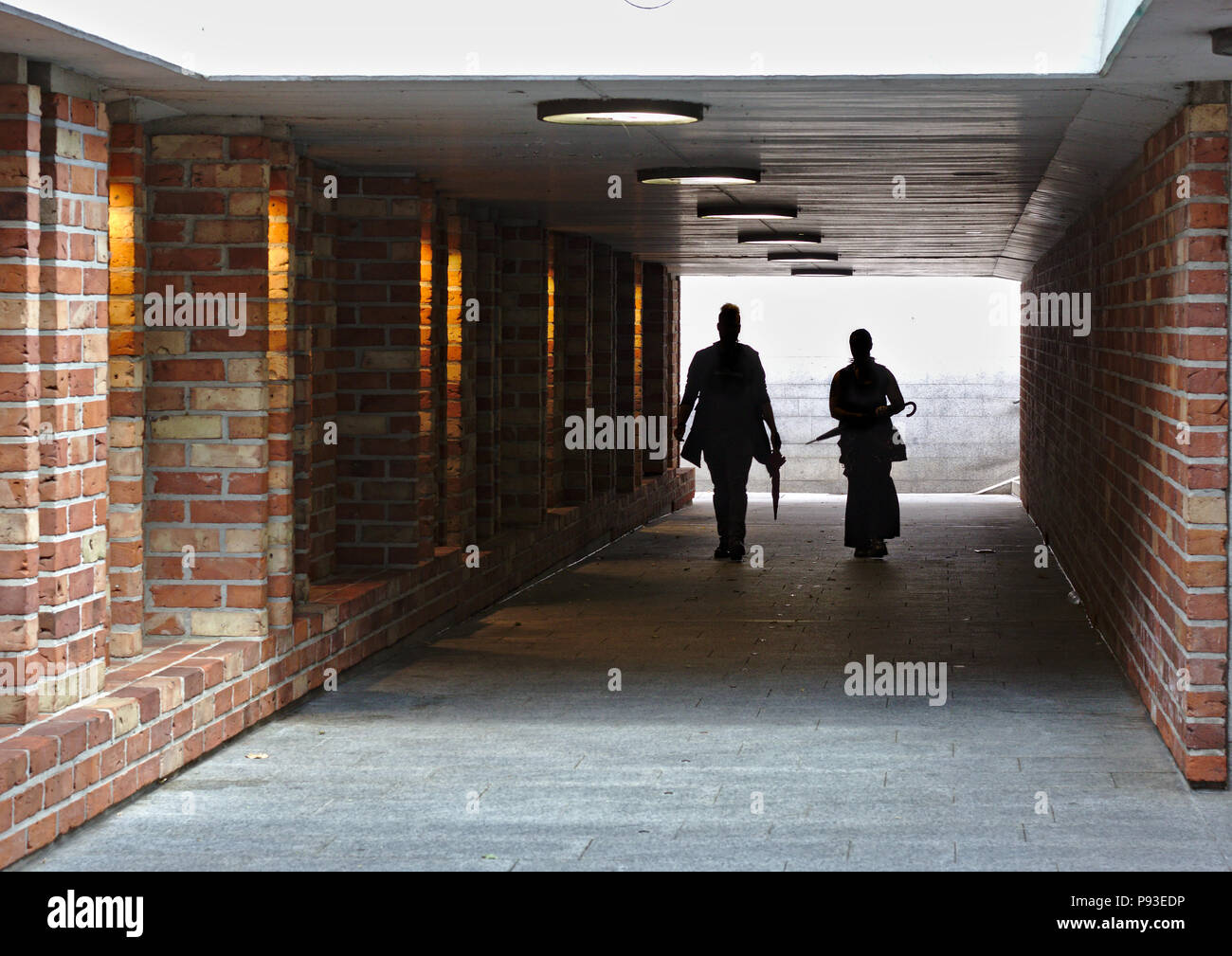 Due persone stagliano contro una luce brillante a piedi attraverso un tunnel con pareti in mattoni verso la telecamera Foto Stock