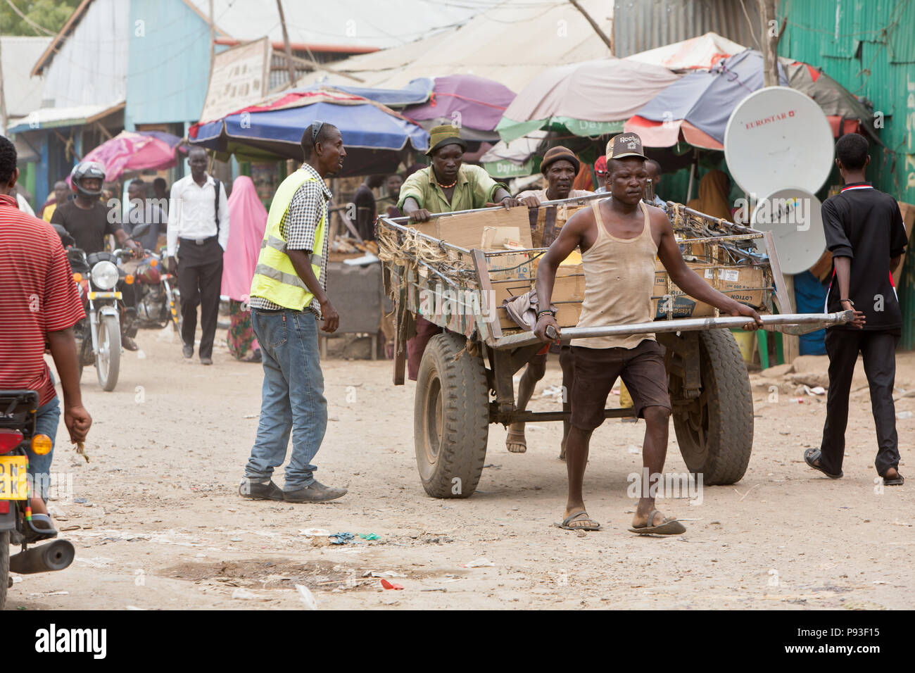 Kakuma, Kenya - Tageloehner tirare un grande rimorchio di trasporto caricata con merci su un occupato, strada sterrata. Foto Stock