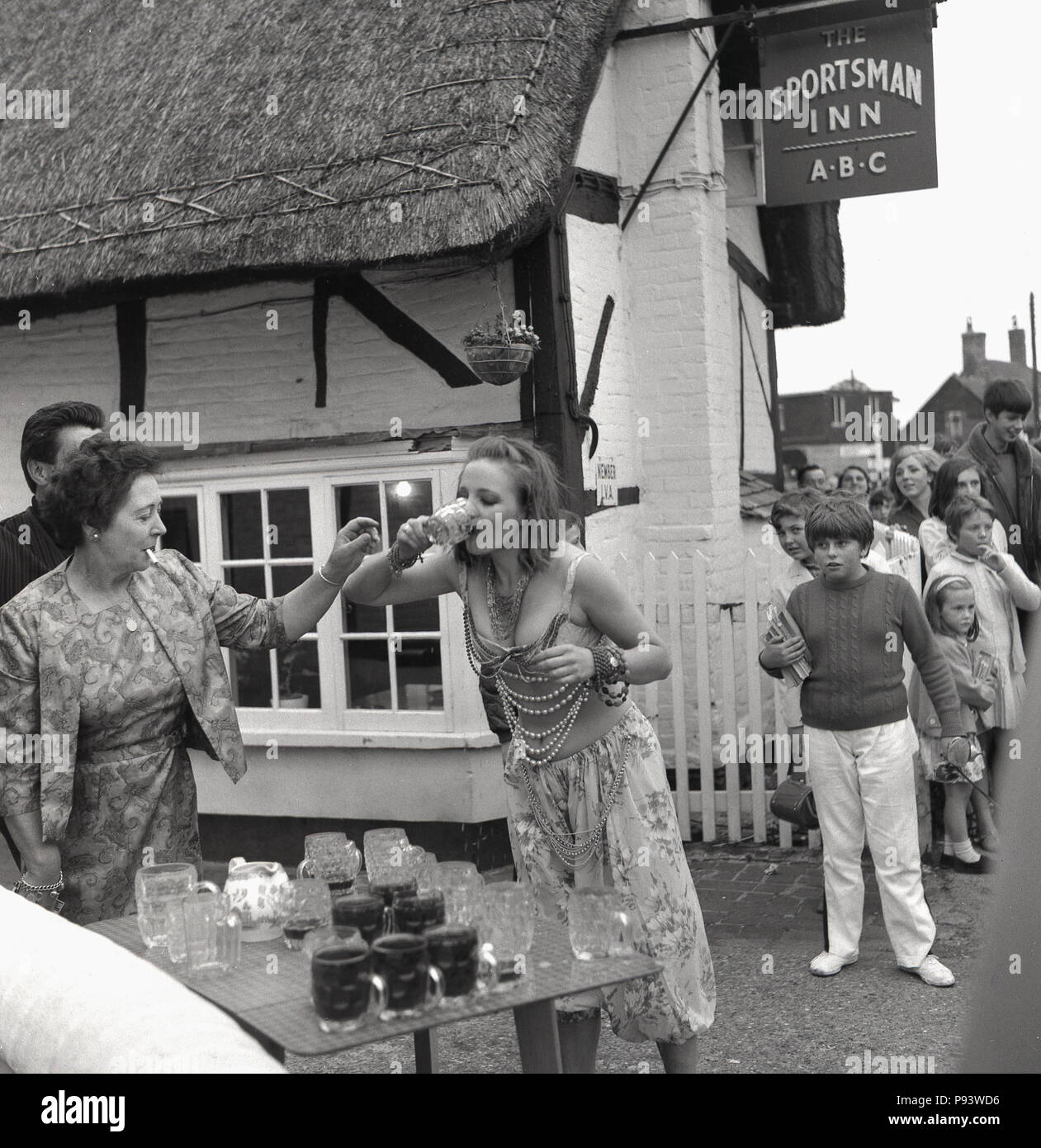 1967, una giovane donna che indossa abiti fantasiosi bevande rapidamente un bicchiere di birra da una tabella di pieni e di vuoti boccali di birra al di fuori lo sportivo Inn at Quainton village, Aylesbury, Bucks, Inghilterra, Regno Unito. Con la FAG in bocca, il ben vestiti padrona di casa è a portata di mano per raccogliere il vetro, mentre un gruppo di giovani stanno a guardare in segno di stupore. La giovane donna è in competizione in un gioco di bere come parte del villaggio fete celebrazioni. Foto Stock