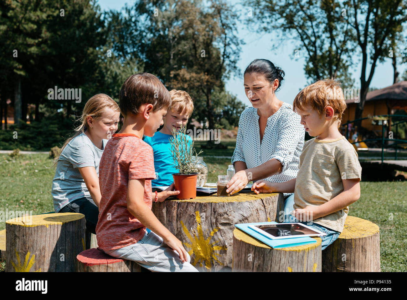 Un gruppo di scolari aventi una lezione al di fuori in un parco. Bambini seduti attorno a un tavolo in legno e imparare insieme con un insegnante in giardino. Foto Stock