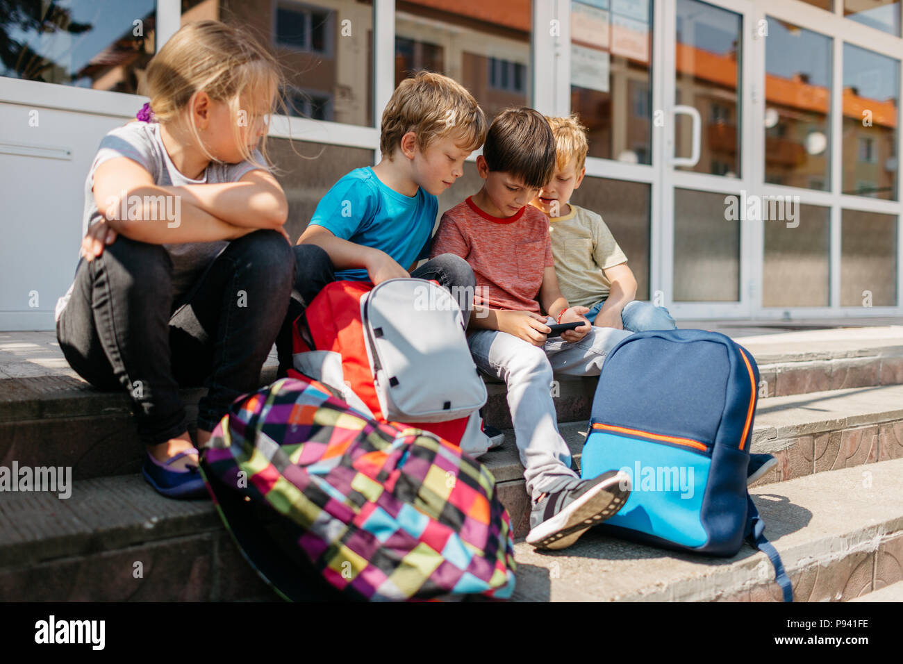 Bambini seduti al di fuori della scuola a giocare con un cellulare. Un gruppo di giovani studenti seduti sulle scale al di fuori della scuola navigando in un telefono. Foto Stock