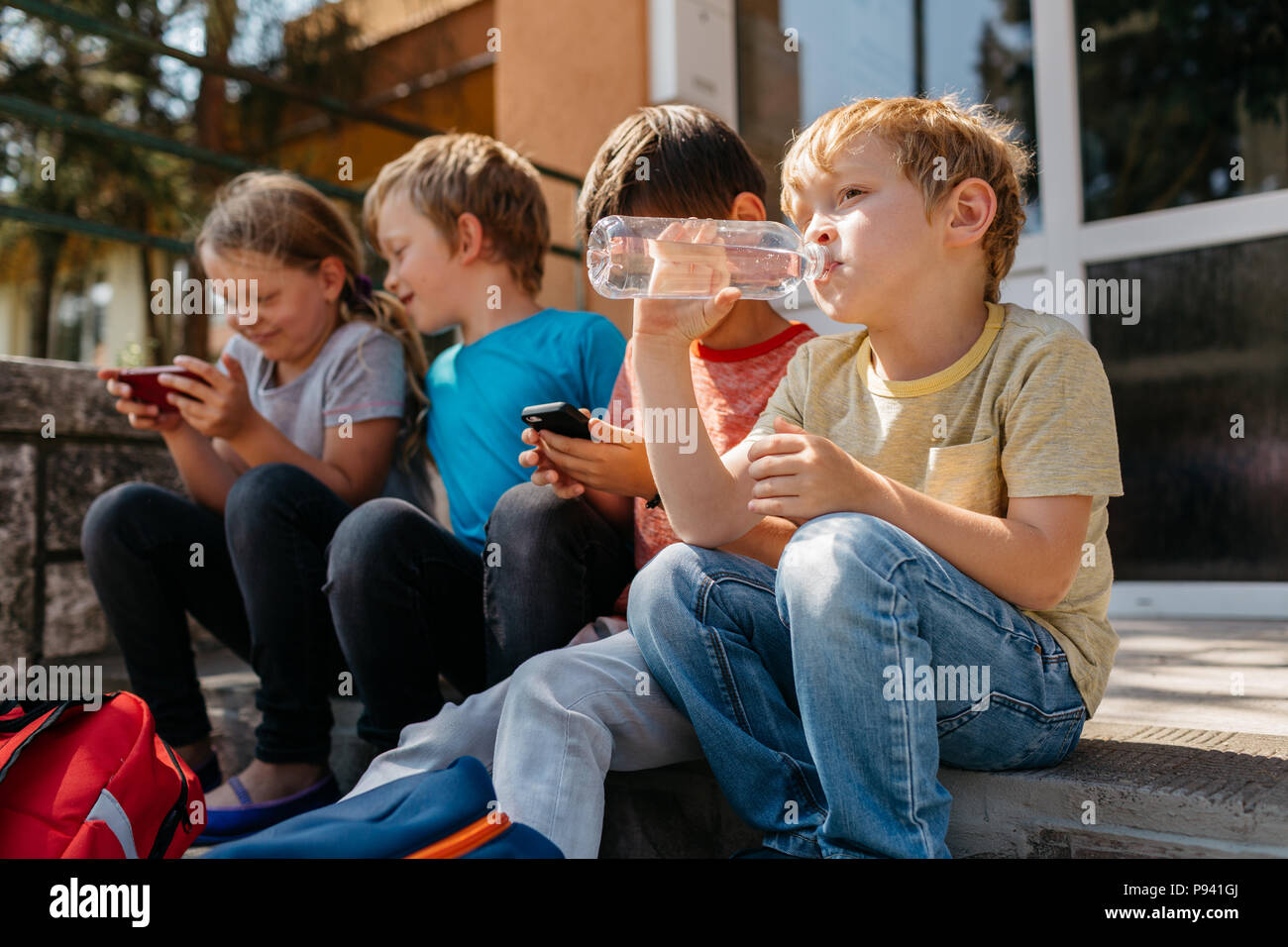 Giovani studenti seduti sulle scale al di fuori della scuola a giocare con il cellulare durante una pausa della scuola. Studenti primari in appoggio di fronte a scuola. Foto Stock