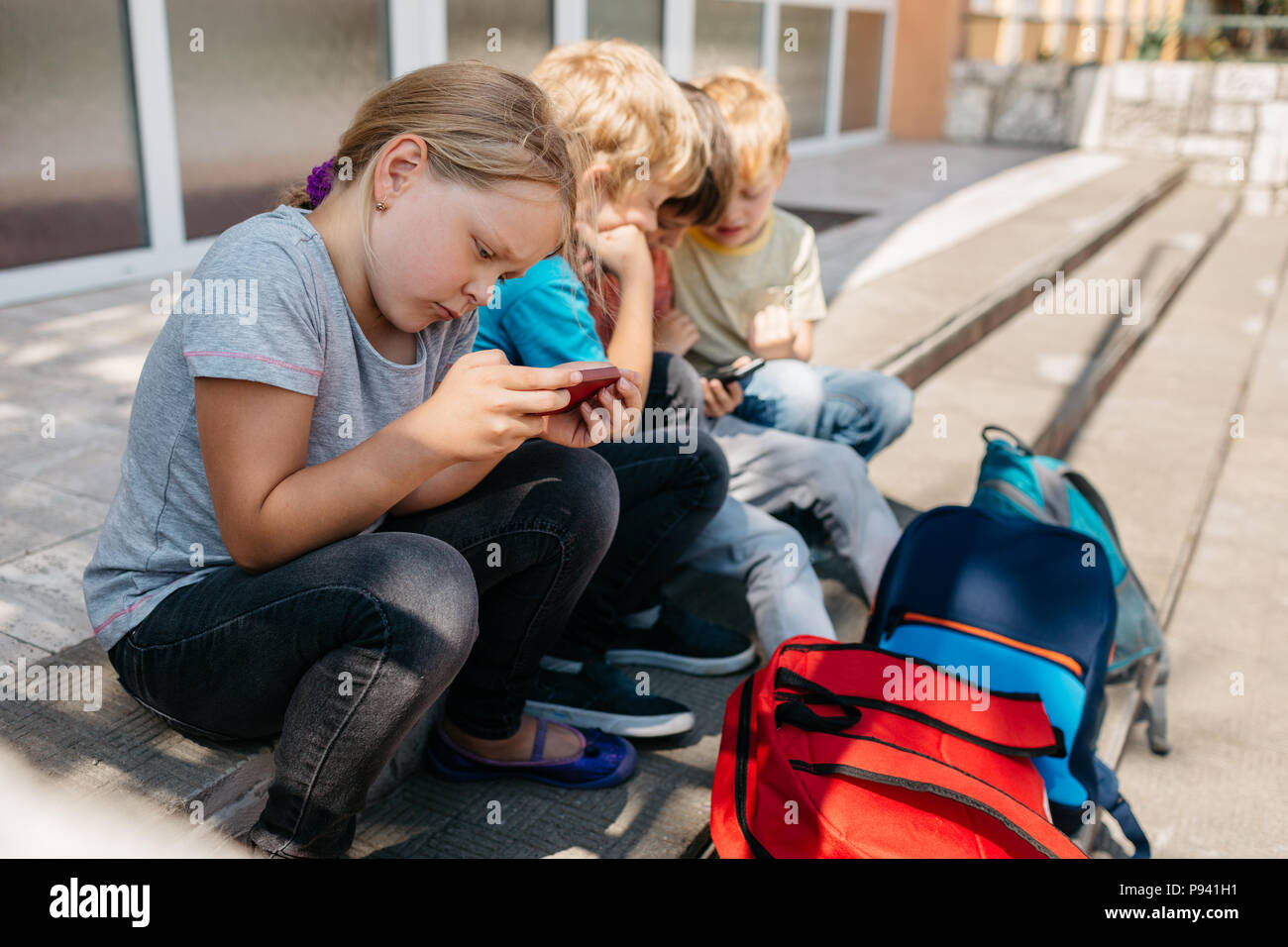 Bambini primaria giocare sui loro telefoni cellulari. Scolari seduti sulle scale durante una pausa della scuola la navigazione. Foto Stock