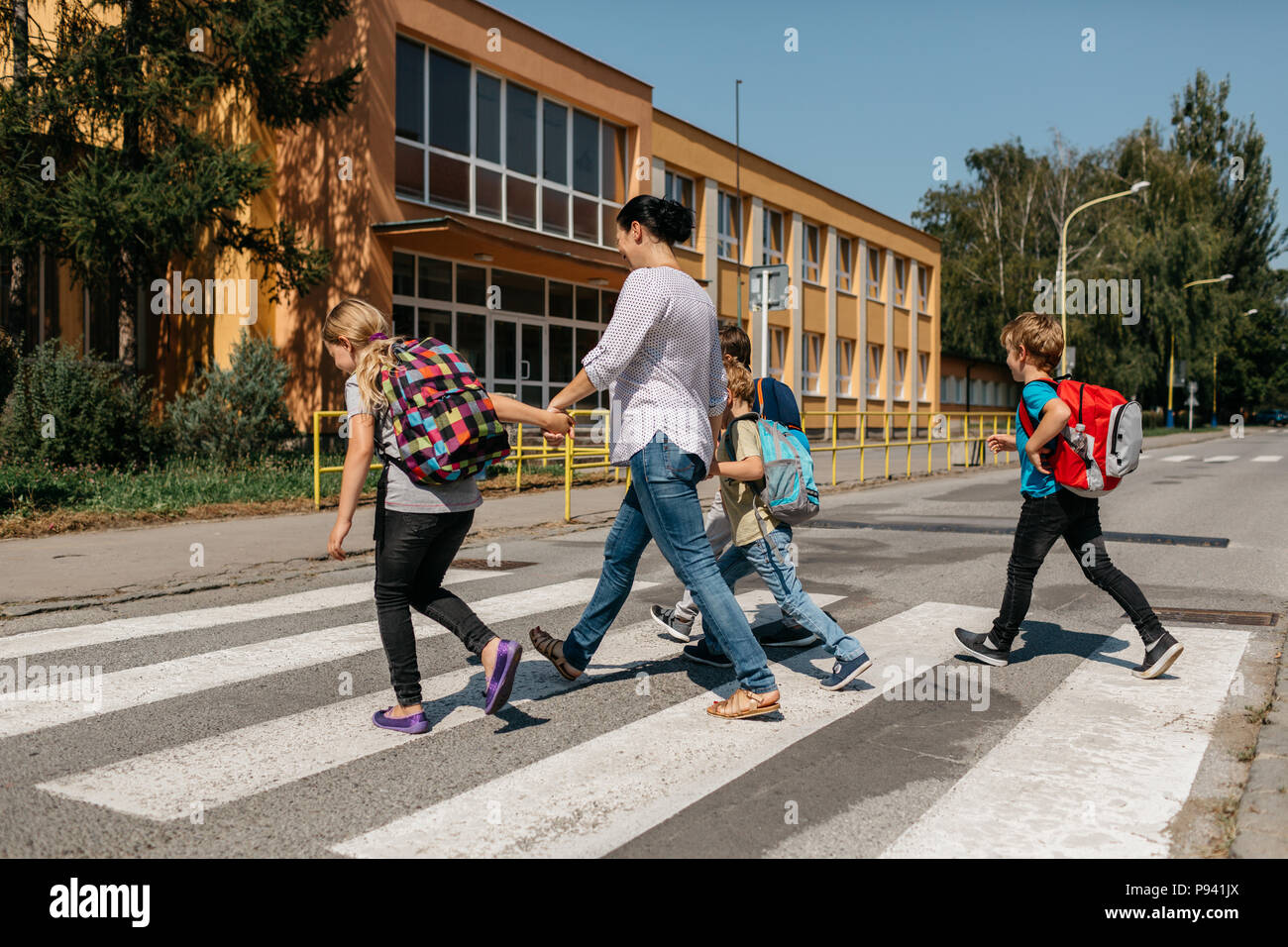 Si torna a scuola - un gruppo di bambini a piedi attraverso una zebra incrocio con un adulto. Vista posteriore dei bambini e la loro madre di attraversare una via. Foto Stock