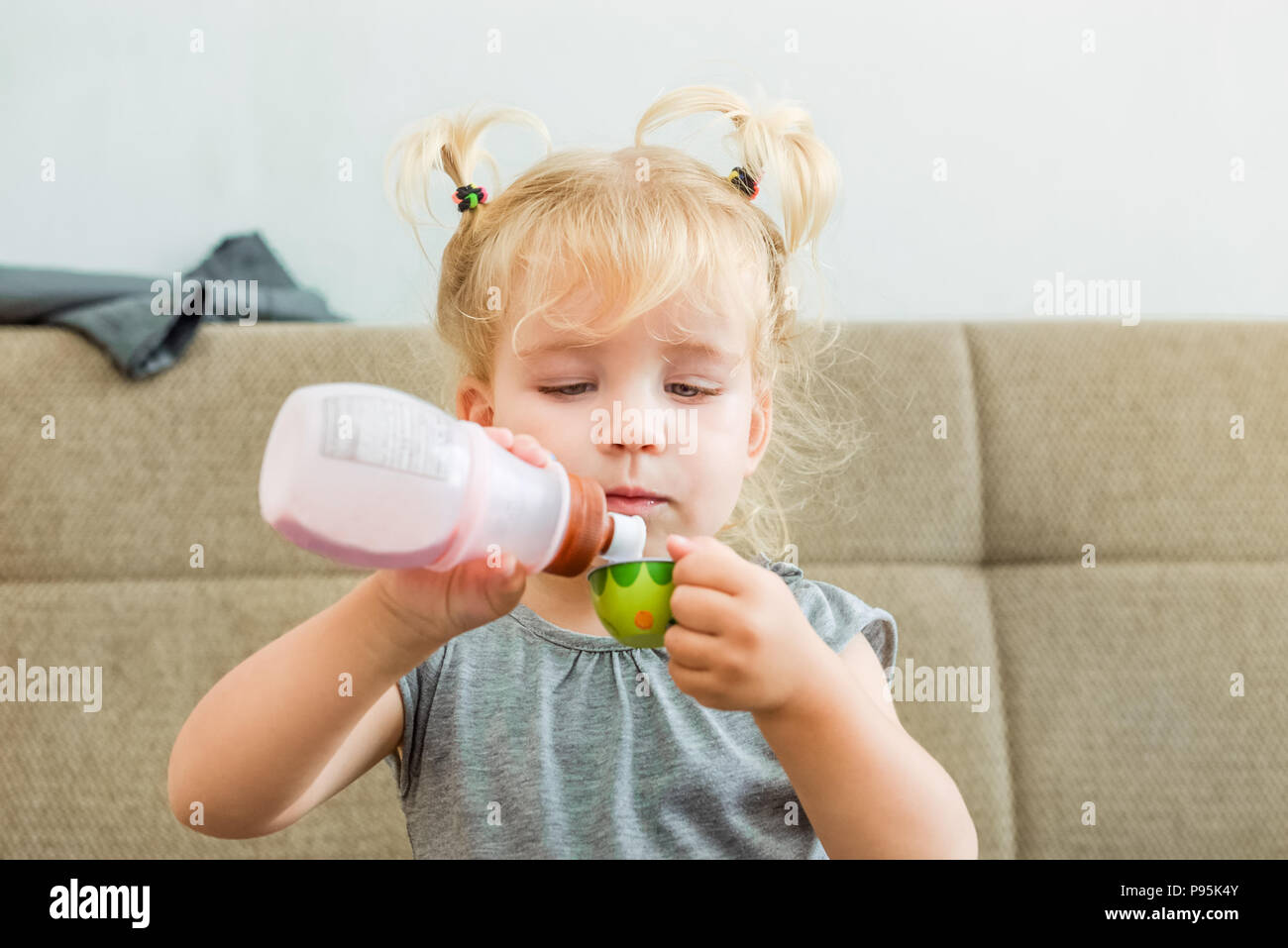 Close up carina ragazza toddler versando lo yogurt naturale dalla bottiglia di plastica a toy cup a casa . Dieta di sane abitudini alimentari. Bambino supporto autonomo, istruzione c Foto Stock