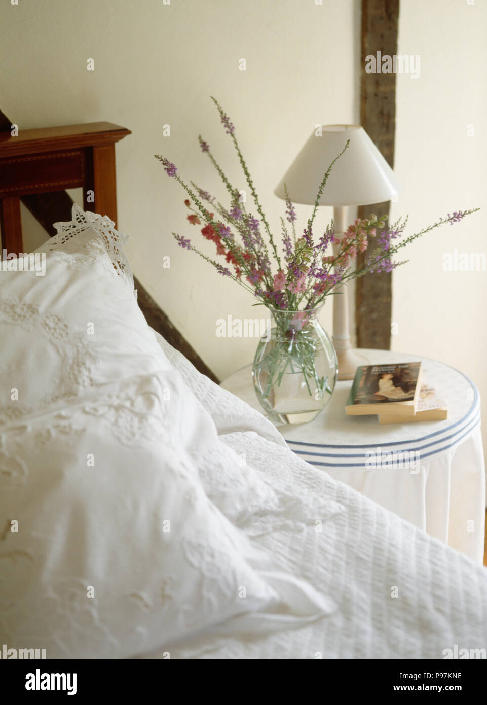 Lampada bianca e fiori freschi in vaso di vetro sul comodino accanto al letto con il bianco broderie anglaise cuscini Foto Stock
