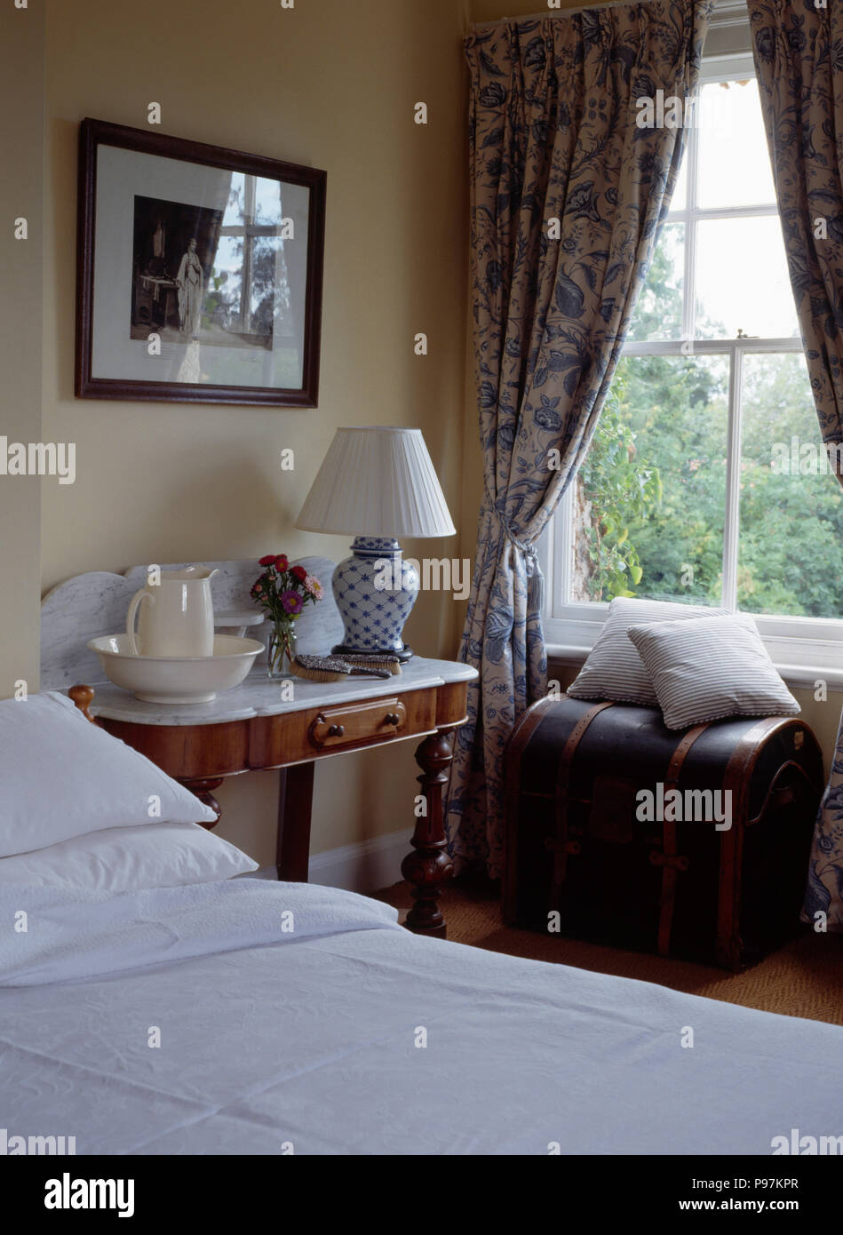 Brocca+ewer sul marmo comodino in camera da letto del paese con blue+tende bianche sulla finestra sopra il torace Foto Stock