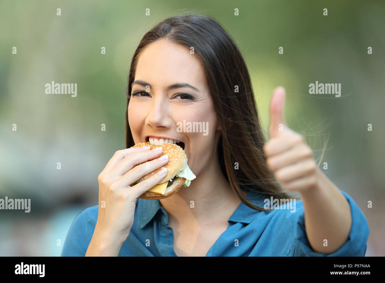 Donna felice mangia un hamburger con il pollice in alto guardando la fotocamera su strada Foto Stock