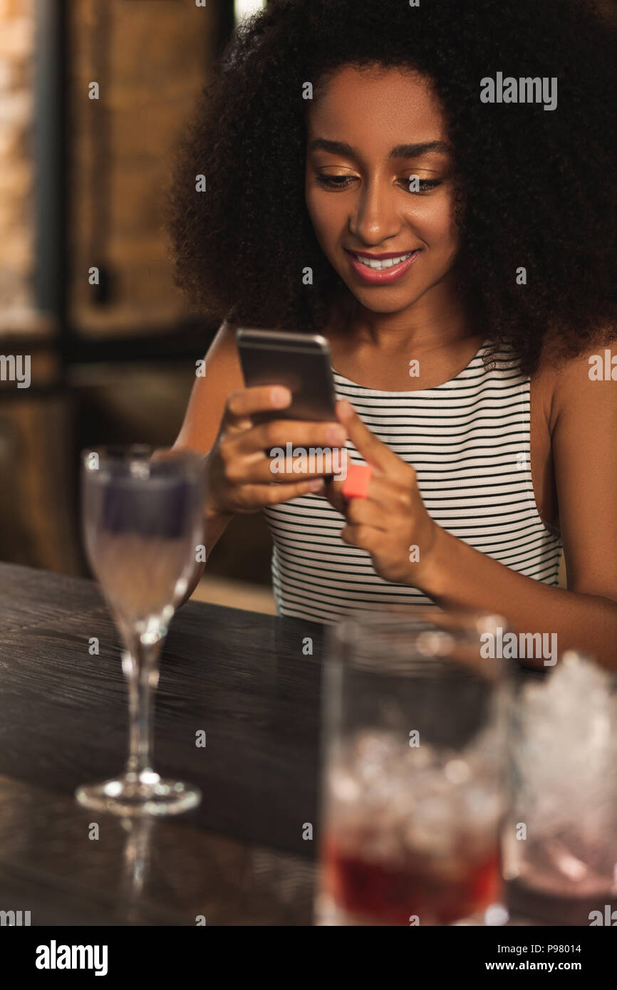 Bella donna ricci texting pur avendo un cocktail nel bar Foto Stock