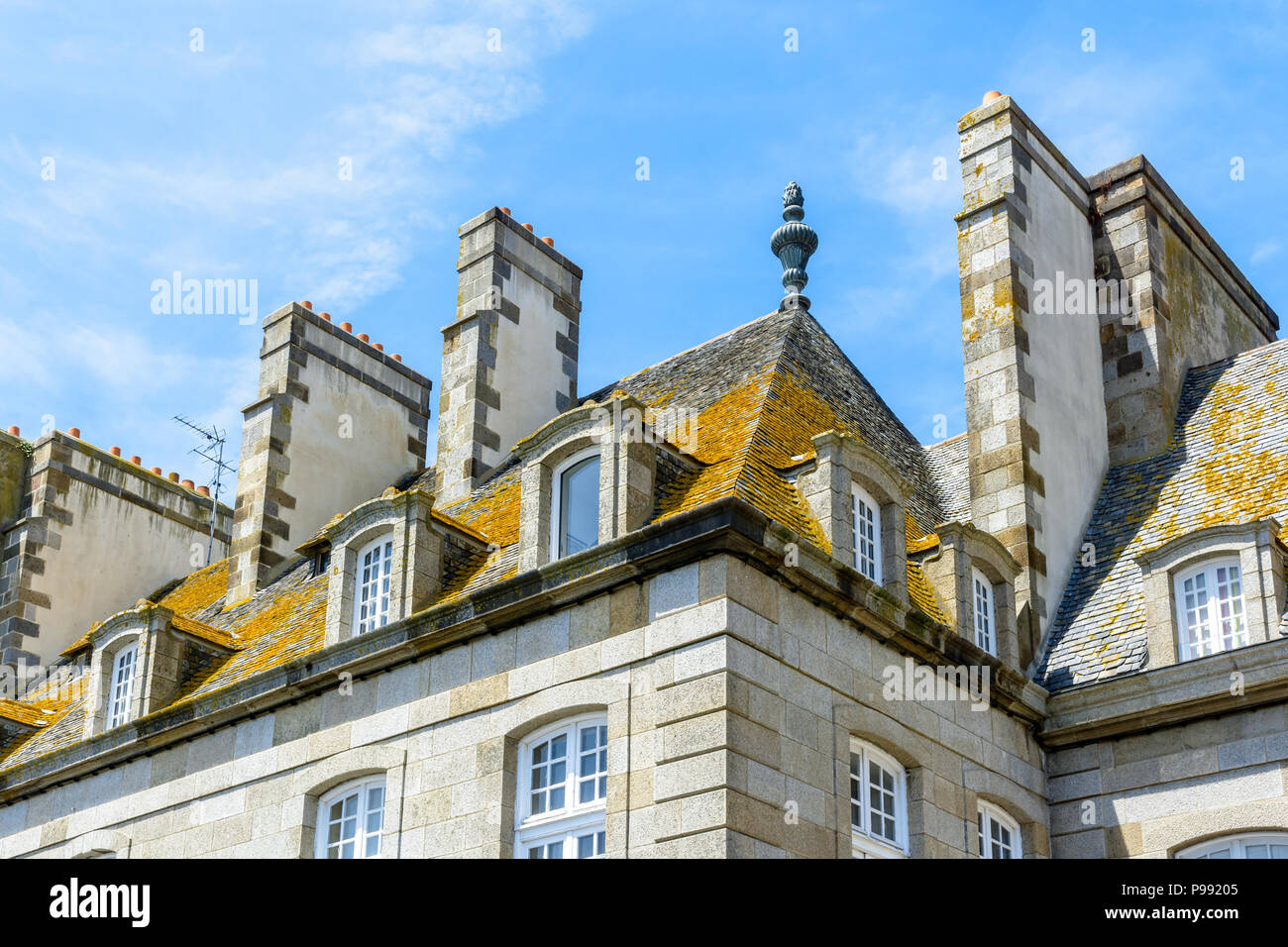 La parte superiore di un edificio residenziale nella città vecchia di Saint-Malo con tetto in ardesia ricoperta di licheni, abbaini e camini contro il cielo blu. Foto Stock