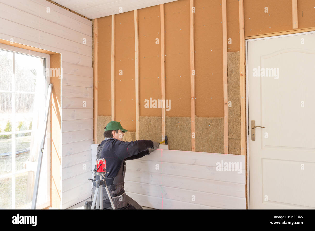 Il lavoratore fa opere di finitura delle pareti con un bianco tavola di legno, utilizzando una linea laser livello. Costruzione di isolante termico eco-telaio in legno casa con legno piastre in fibra Foto Stock