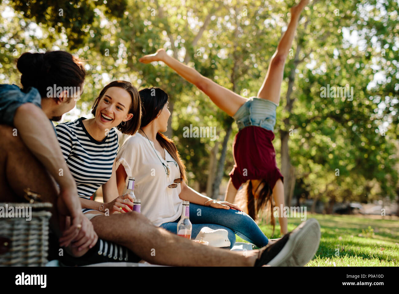 Gruppo di amici seduti al parco con una donna facendo un salto appoggiate in background. Giovani uomini e donne godendo di un giorno di estate al parco. Amici enjoyin Foto Stock