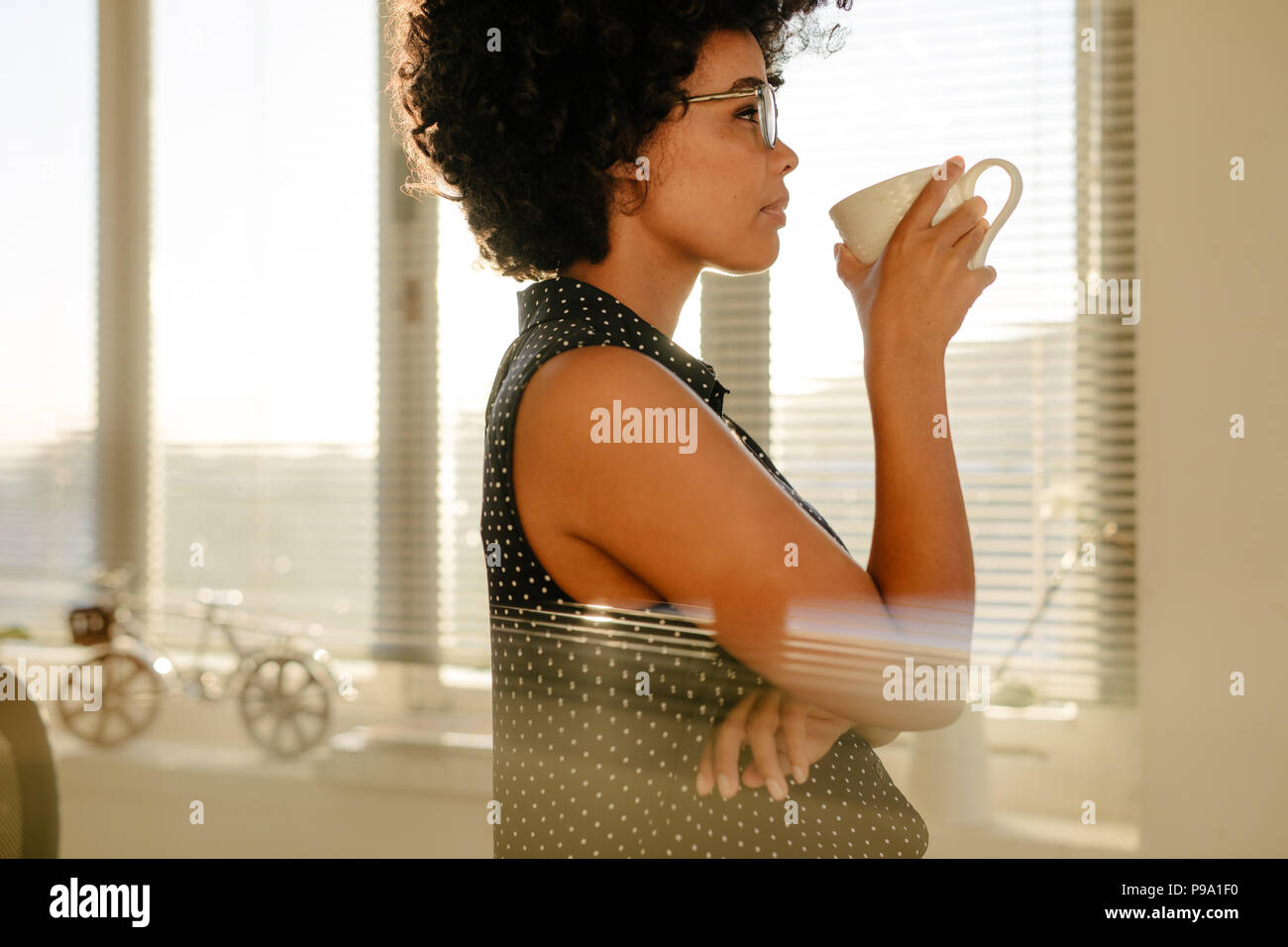 Vista laterale del giovane imprenditrice in casuals di bere il caffè in ufficio. Femmina pensando a qualcosa mentre un caffè durante la pausa di lavoro. Foto Stock