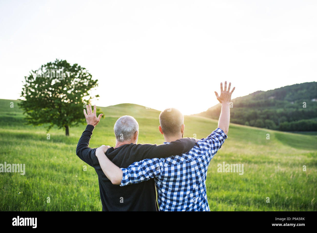 Un adulto hipster figlio col padre senior in una passeggiata nella natura al tramonto, le braccia intorno all'altro. Foto Stock