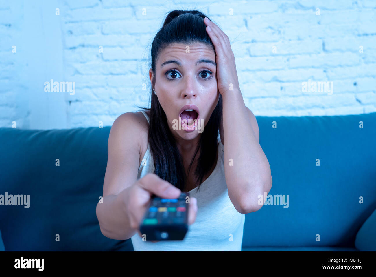 Giovane bella donna latina seduti a casa divano divano in soggiorno a guardare la televisione scary horror movie o orribili notizie spaventato eccitato e il coperchio Foto Stock