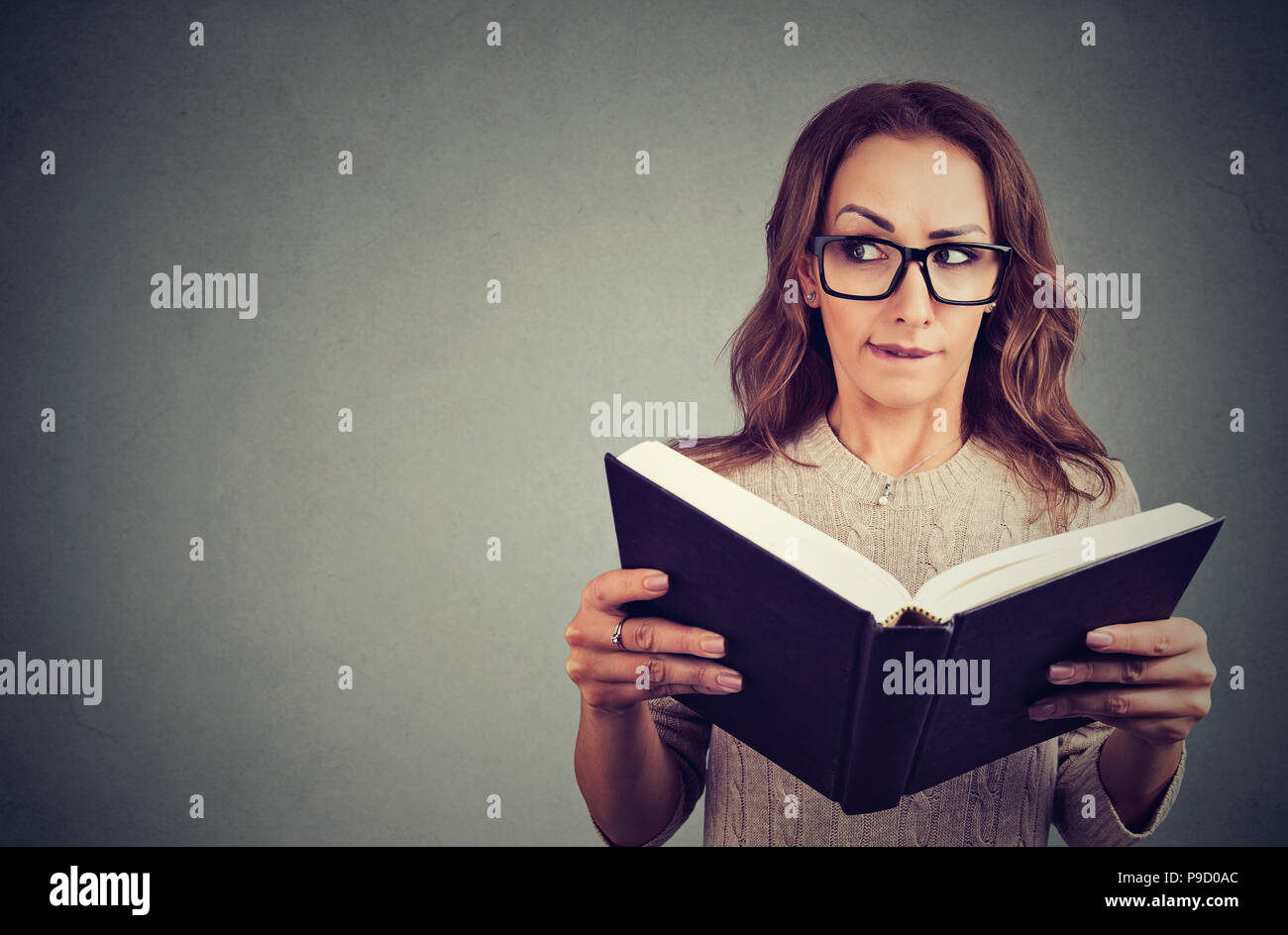 Giovane donna curiosa in bicchieri guardando lontano tenendo premuto e la lettura di libro con storie segrete su sfondo grigio Foto Stock