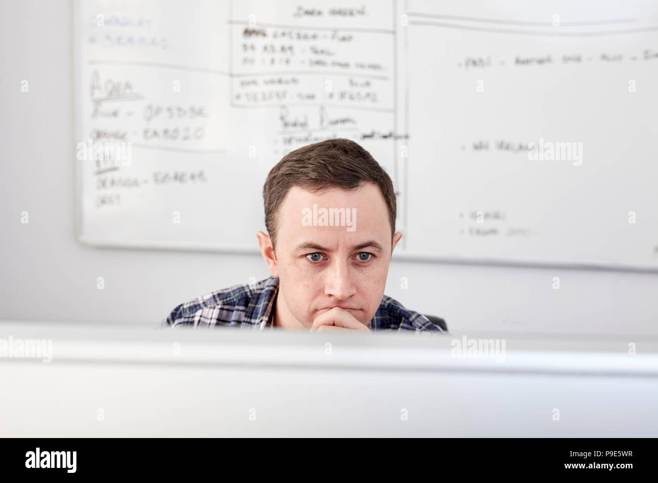 Uomo che guarda uno schermo di computer seduto alla sua scrivania, con un progetto grafico sulla lavagna dietro. Foto Stock