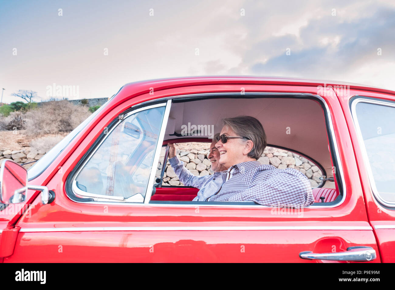 Coppia di anziani con cappello, con occhiali, con il grigio e il bianco dei capelli, con camicia casual, su vintage auto rossa in vacanza godendosi il tempo e la vita. Con un chee Foto Stock