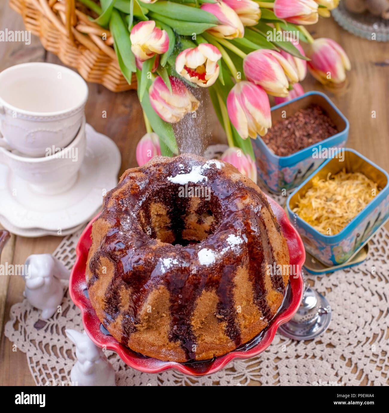 In casa torta dolce, versata con cioccolato, su una torta rossa piastra. Su uno sfondo di legno secco, erbe per tè e tazze. Un mazzo di fiori di primavera. Tulipani e un coniglio di ceramica. Cartolina e spazio libero Foto Stock