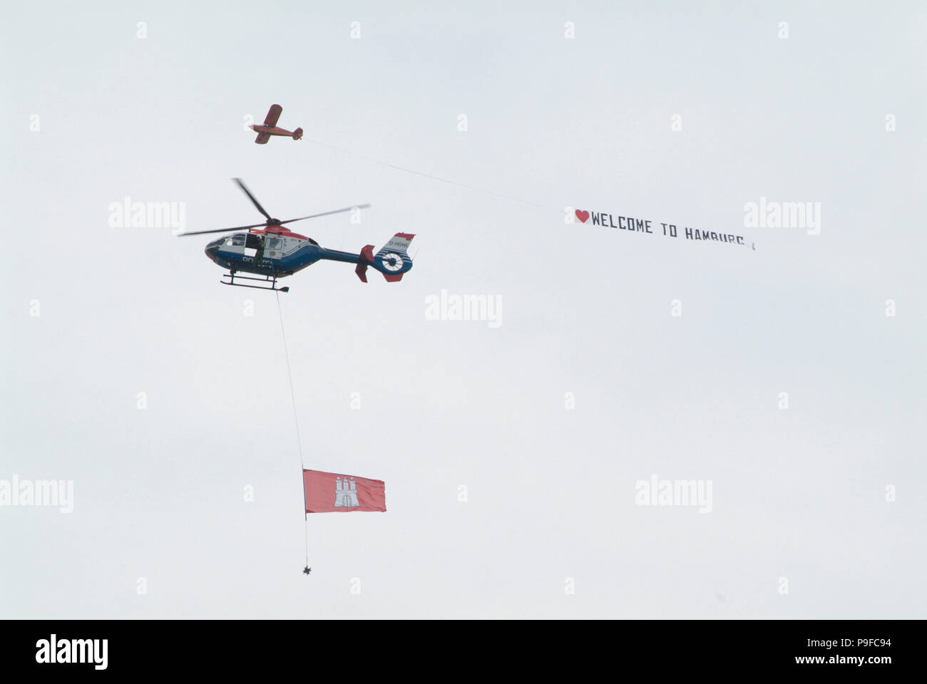 Einmotoriges Flugzeug zieht ein Werbebanner über den Hamburger Hafen: benvenuti ad Amburgo. Davor ein Polizei-Hubschrauber mit großer Hamburg-Flagge Foto Stock
