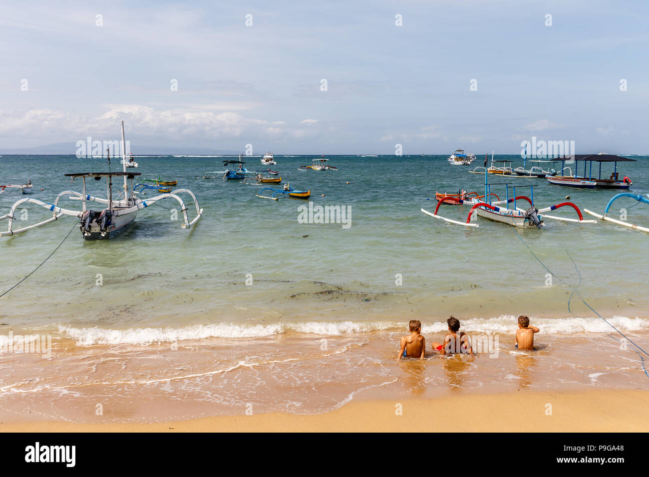 Indonesiano tradizionale stile outrigger legno barche da pesca (jukung) e albero kids in seduta la marea sulla spiaggia di Sanur, Bali, Indonesia. Foto Stock