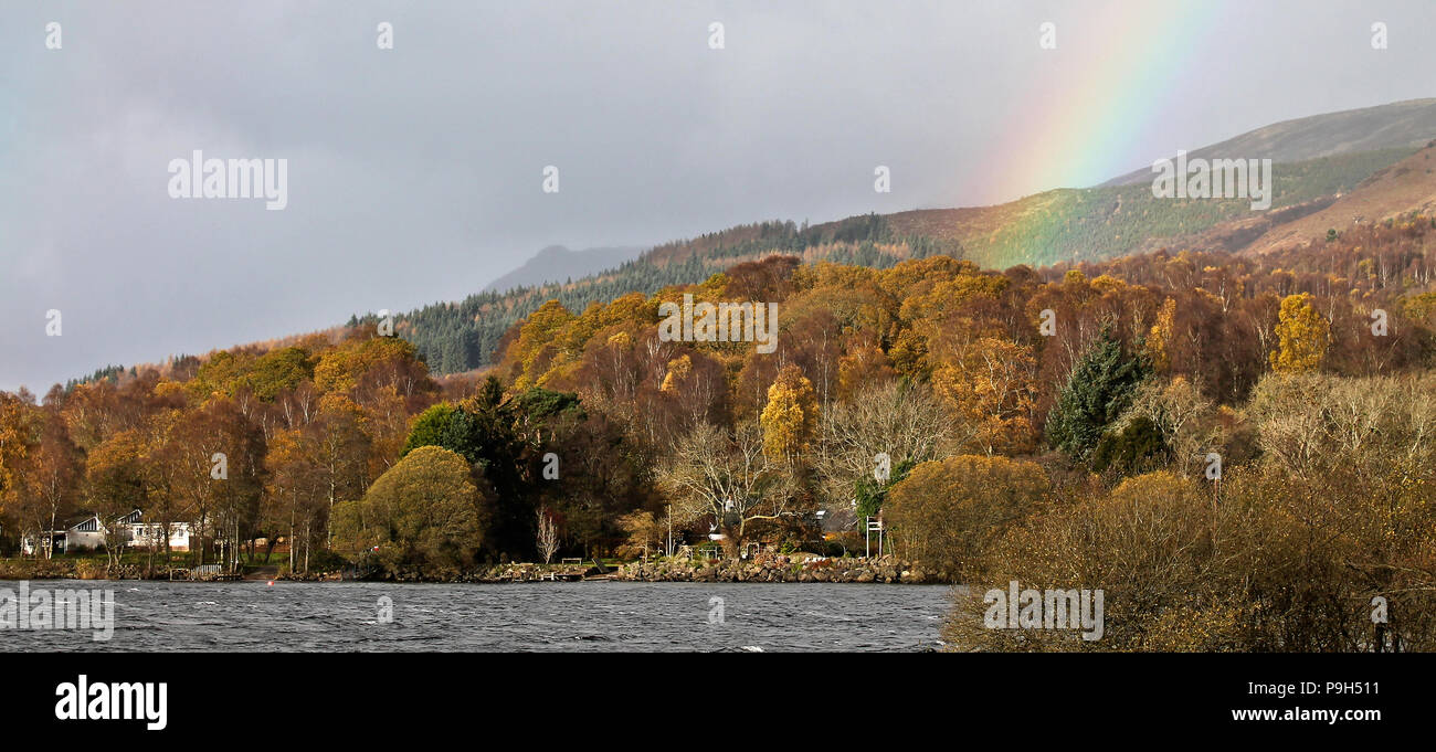 Le foglie di autunno sugli alberi e montagne che sorgono dalla sponda orientale del Loch Lomond in Scozia con un arcobaleno aggiunta alla scena. Foto Stock