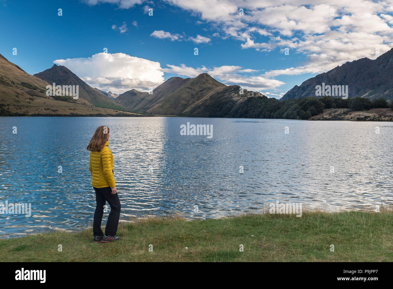 Escursionista femmina in piedi sulla riva del lago fuma vicino a Queenstown, il lago con le montagne, Otago, Isola del Sud, Nuova Zelanda Foto Stock