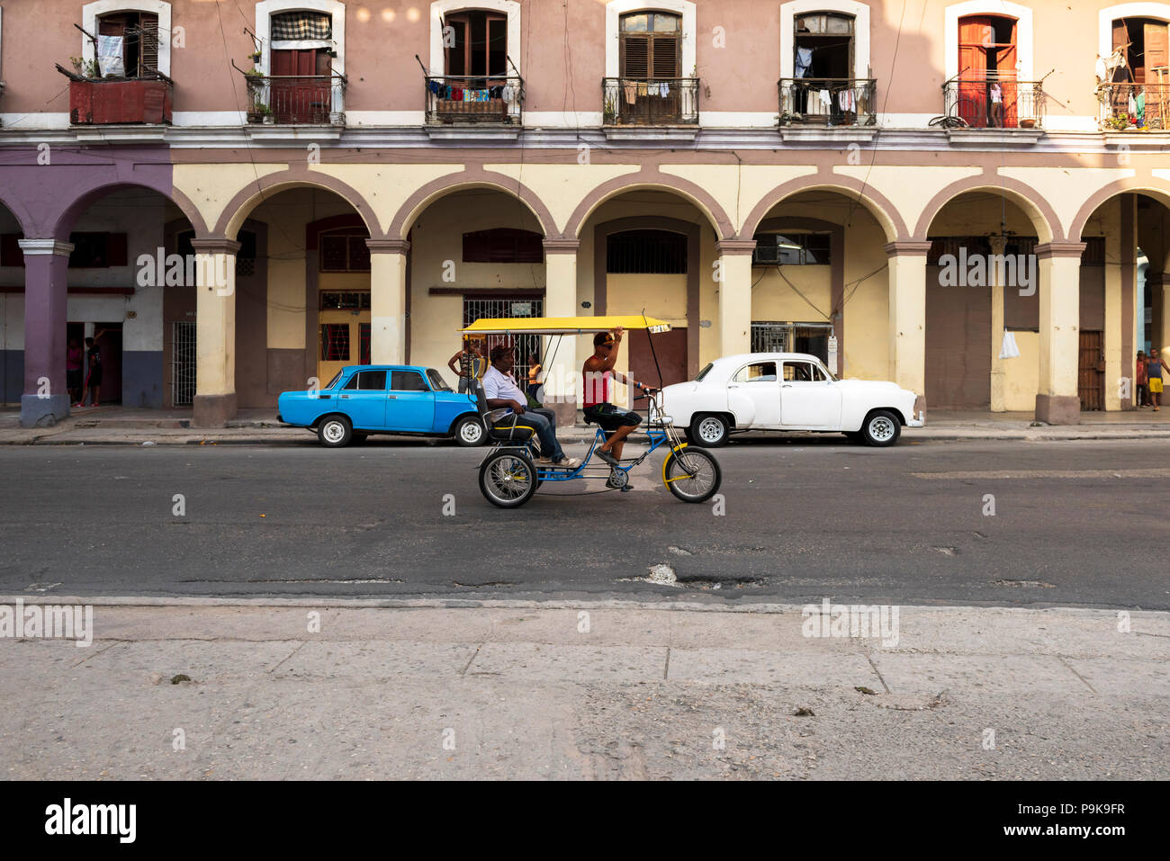 Uomo alla guida di pedicab con passeggero in primo piano; Lada blu e bianco Volga vecchie auto parcheggiata su strada a l'Avana, Cuba. Foto Stock