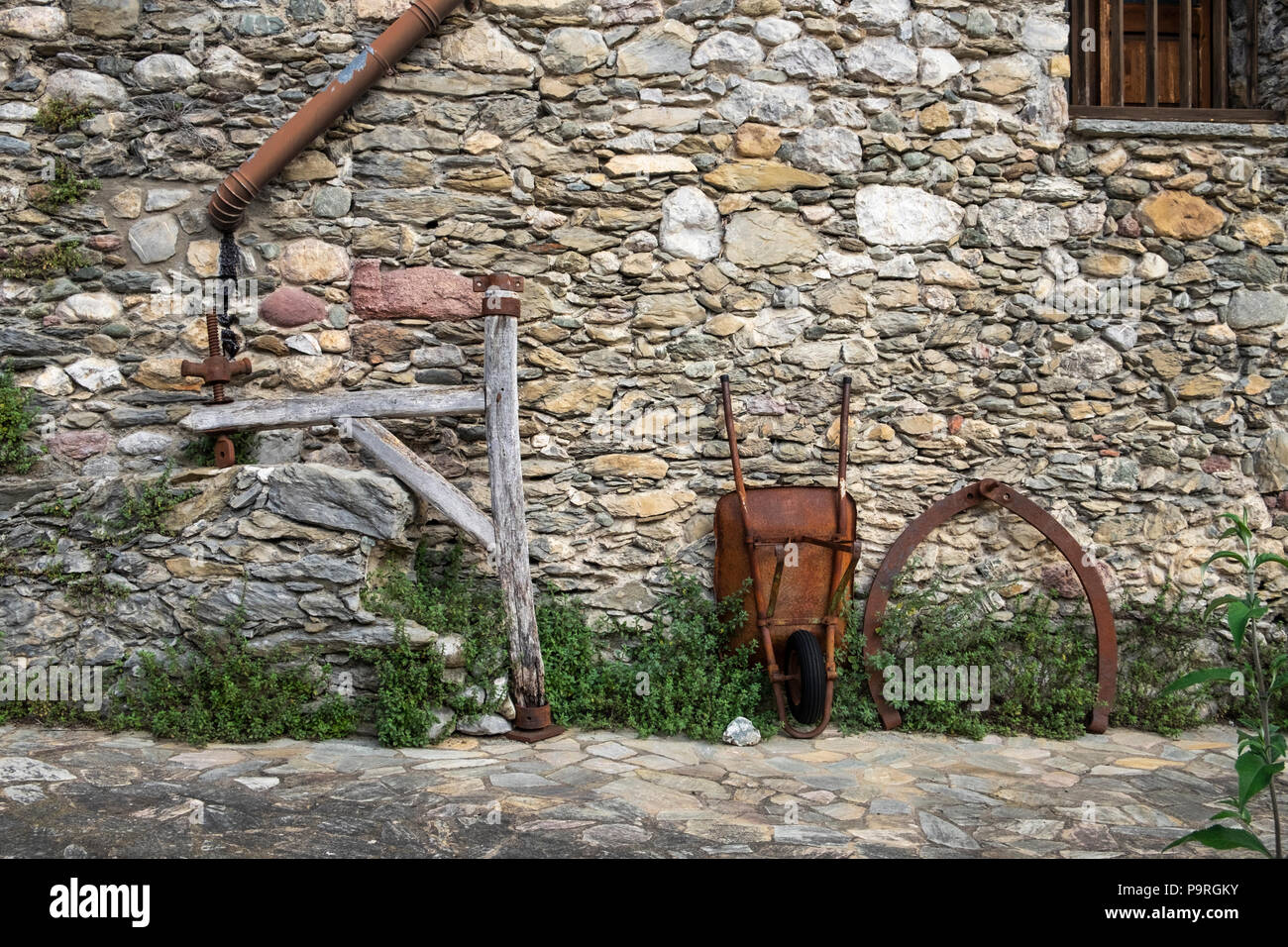 Rusty carriola e altri attrezzi agricoli appoggiata contro una pietra in granito muro di un edificio in generano, Catalogna, Spagna Foto Stock