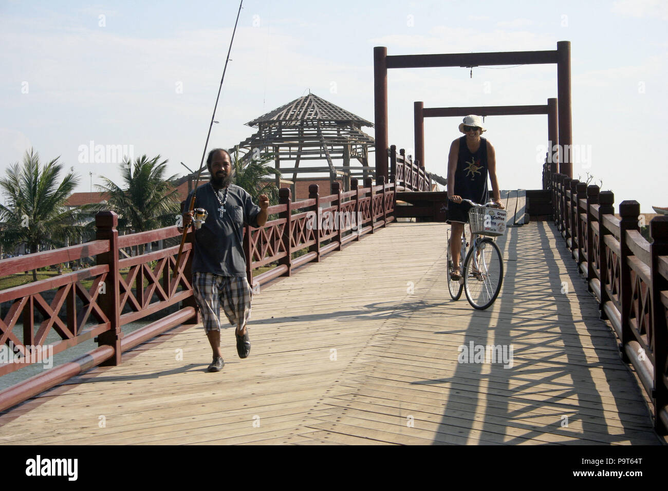 Uomo vietnamita con canna da pesca a piedi attraverso un ponte con turisti in bicicletta dietro, vicino a Mui Ne, Vietnam Foto Stock