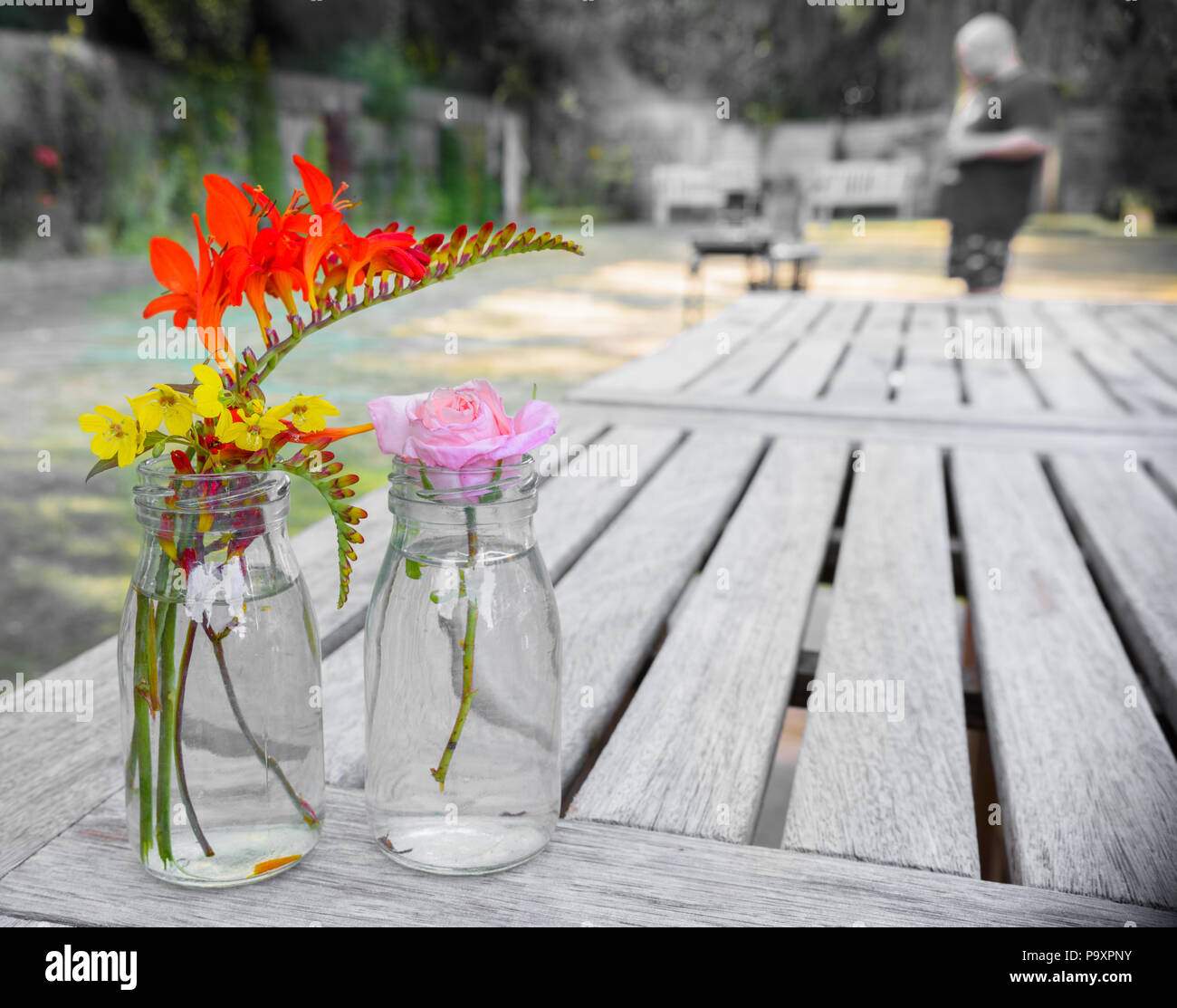 Un paio di vasi di fiori colorati su un tavolo di legno in un giardino inglese su una soleggiata giornata estiva, con un uomo in piedi su un barbecue in backgro Foto Stock