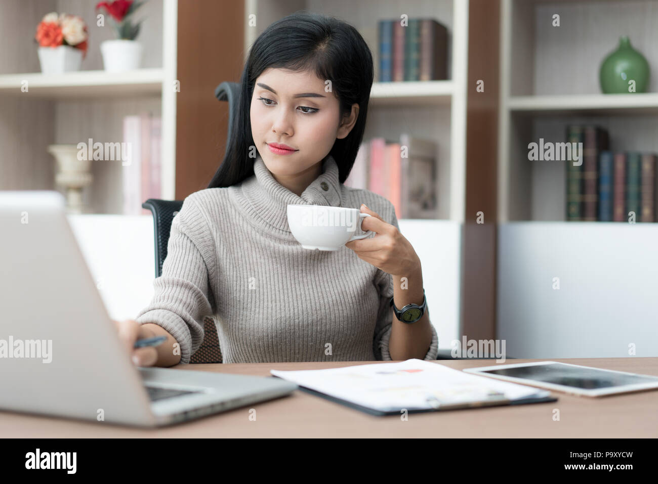 Giovani asiatici imprenditrice seduta in ufficio business alla scrivania, a bere caffè e utilizzando laptop. Sul tavolo è portatile. imprenditrice navigando su internet, c Foto Stock