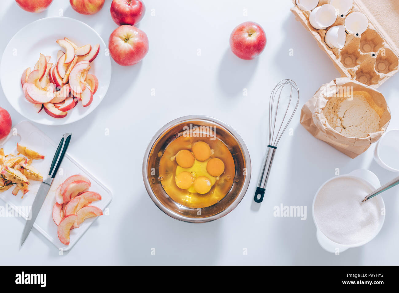 I componenti e gli utensili per la cottura della torta di mele, vista dall'alto. Recipiente metallico riempito con materie tuorli ed albumi vicino a frusta, tagliare la frutta, gusci d'uovo, farina e s Foto Stock