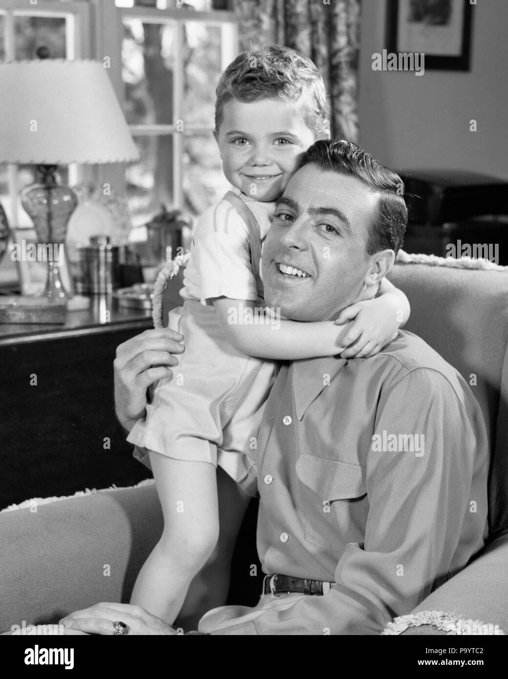 Anni Cinquanta uomo sorridente in POLTRONA CON TODDLER Figlio abbraccia Lui attorno al collo guardando telecamera - J3909 HAR001 HARS il novellame di stile per il viso equilibrio di sicurezza collo abbraccio forte figli lieta gioia LIFESTYLE HOME vita di metà lunghezza Hug persone ispirazione caring maschi abbracciando espressioni padri B&W contatto visivo stretto allegro forza papà orgoglio sorride il collegamento facile gioiosa elegante supporto attacco personale affetto emozione la crescita di lui il novellame relax convivere in bianco e nero di etnia caucasica HAR001 in vecchio stile Foto Stock